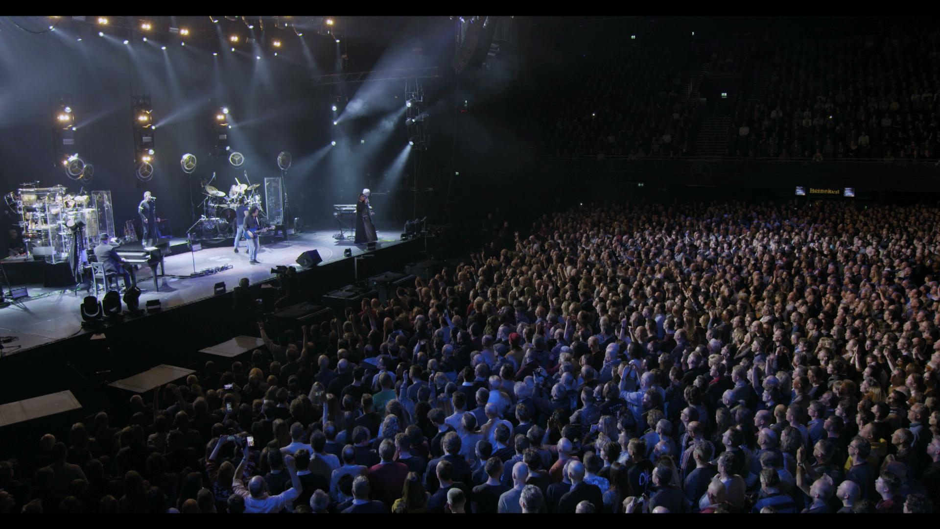 A large crowd watches totoon a well-lit stage with multiple musicians in a concert hall.