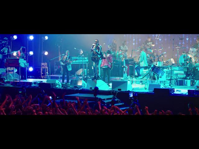 Arcade Fire performs on stage with various instruments. Blue and purple lighting illuminates the scene. An enthusiastic crowd with hands raised is visible in the foreground.