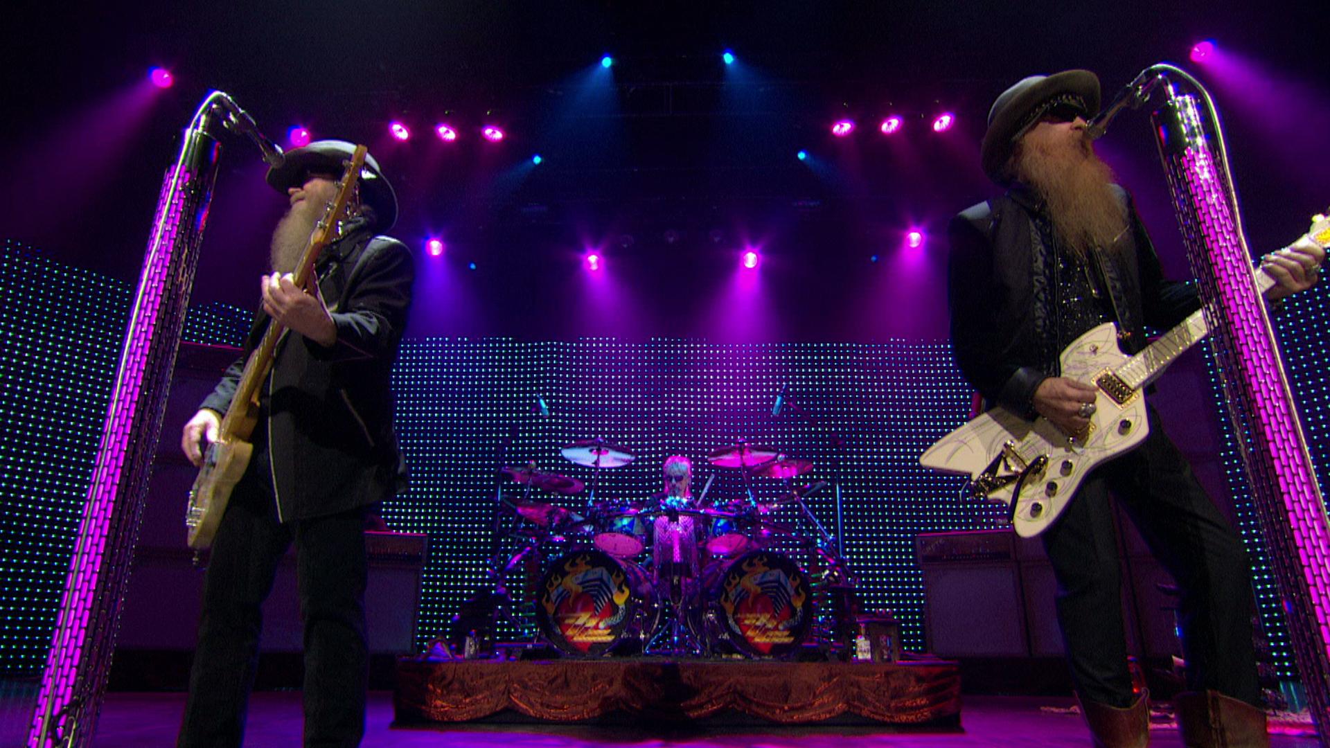 ZZ Top performing on stage with guitars and drums, illuminated by purple and pink stage lights.