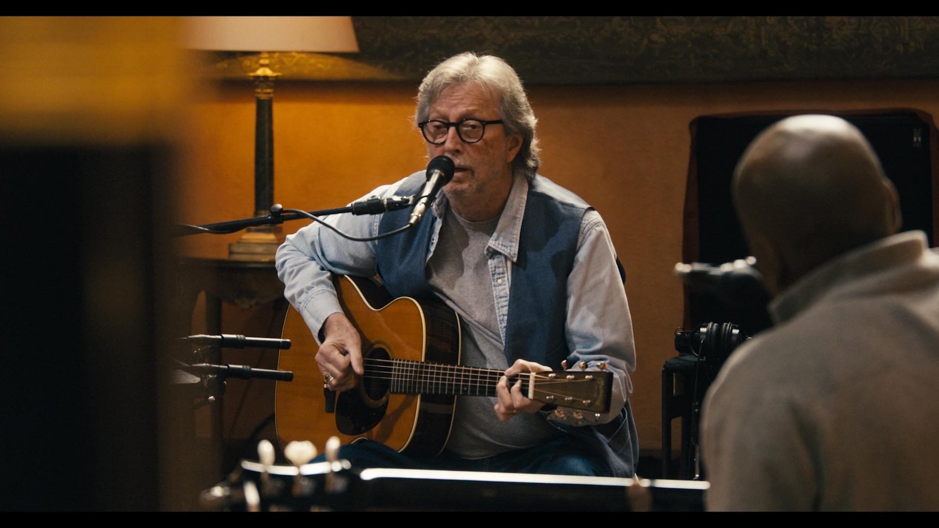 Eric Clapton wearing glasses and headphones plays an electric guitar in a recording studio, with a double bass partially visible in the foreground.