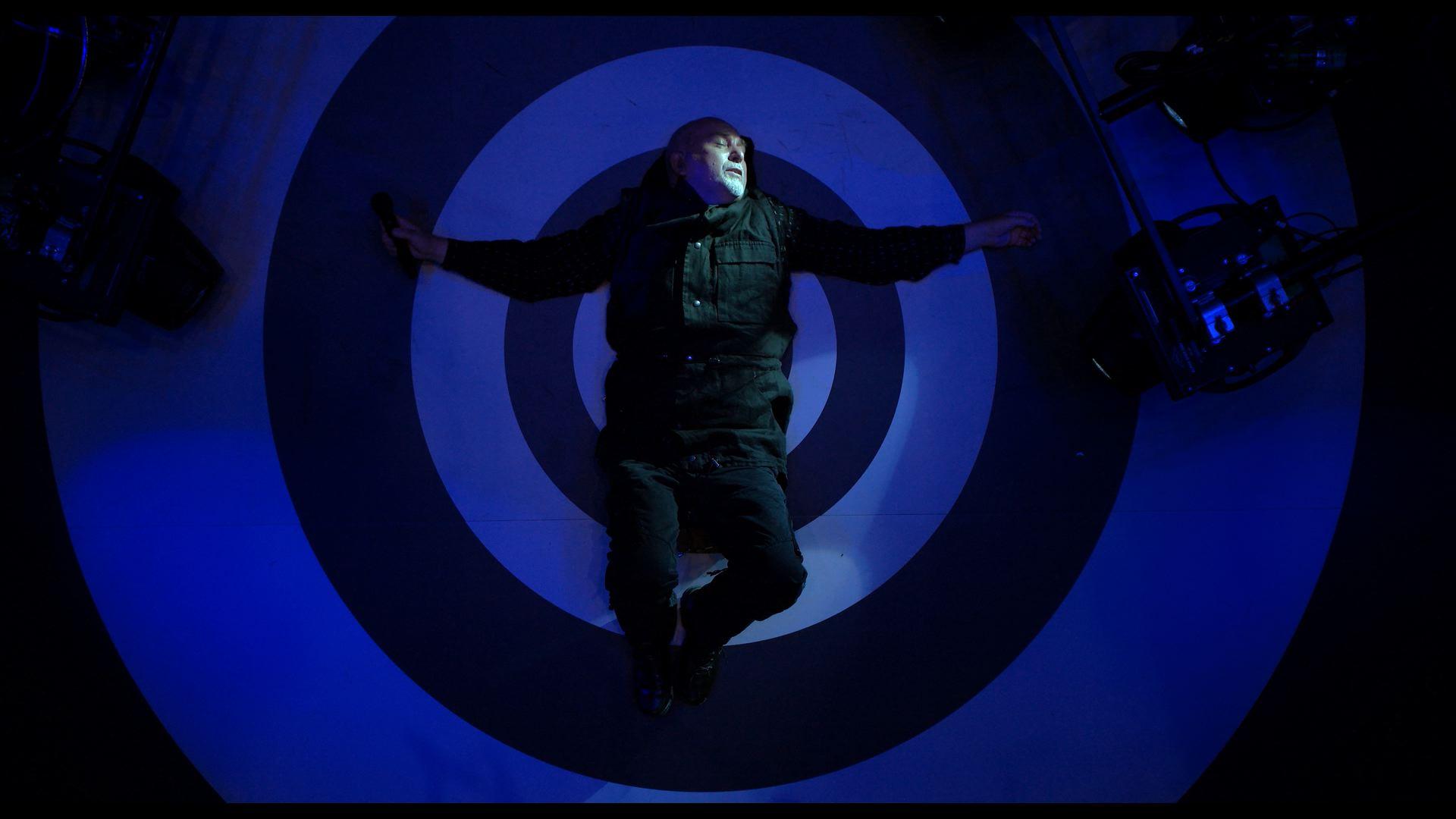 Peter Gabriel lies on their back with arms outstretched in the center of a circular blue and white target design on the floor, surrounded by equipment.