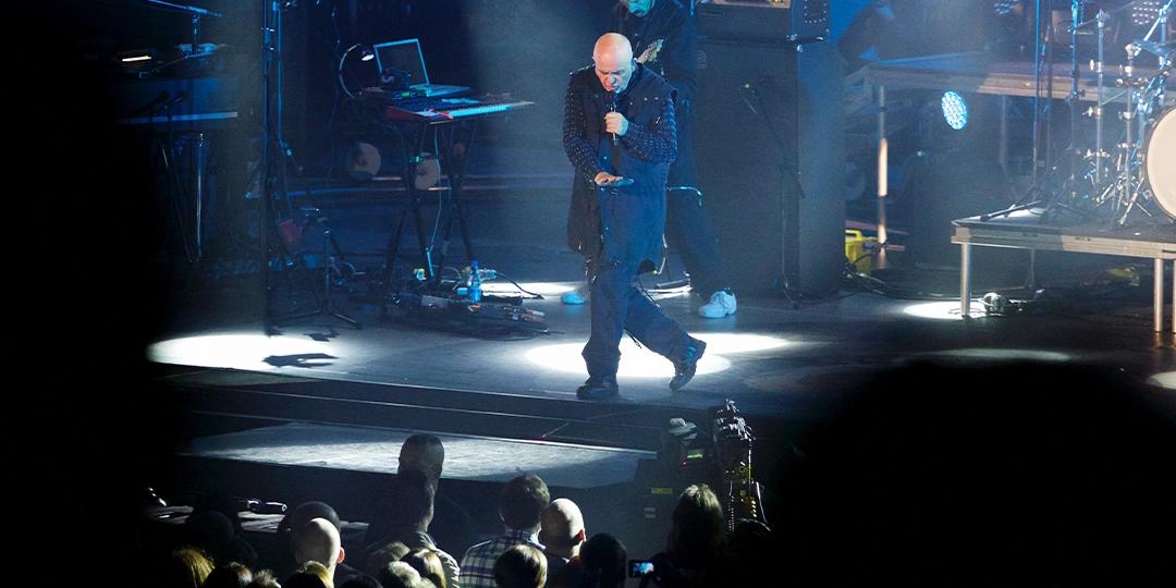 Peter Gabriel with a microphone performs on a dimly lit stage, surrounded by musical instruments and equipment, while an audience watches.