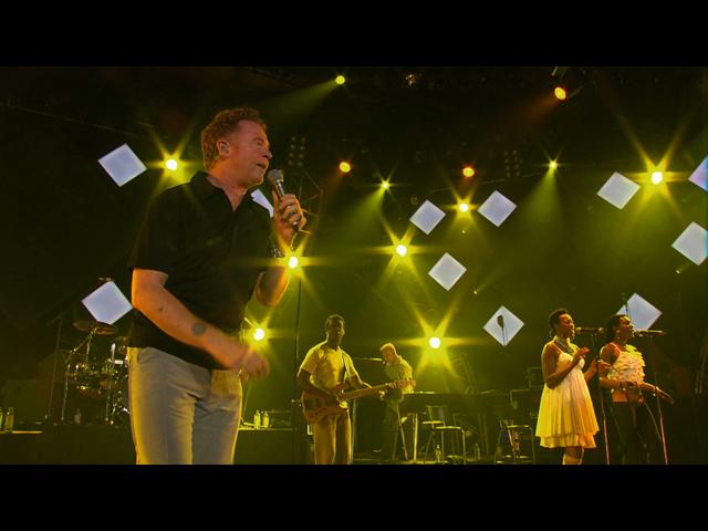 Simply Red performs on a stage with bright yellow lights. The lead singer holds a microphone and stands in front, while the other band members play instruments and sing into microphones.