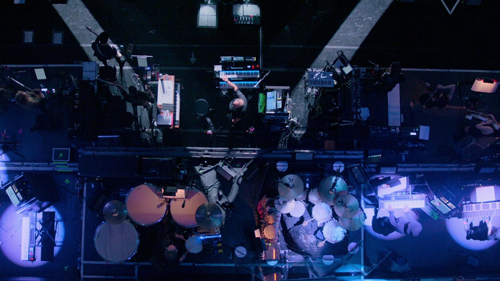 Overhead view of a hans zimmer with various instruments and equipment. Multiple musicians are seen playing drums, keyboards, and other instruments.