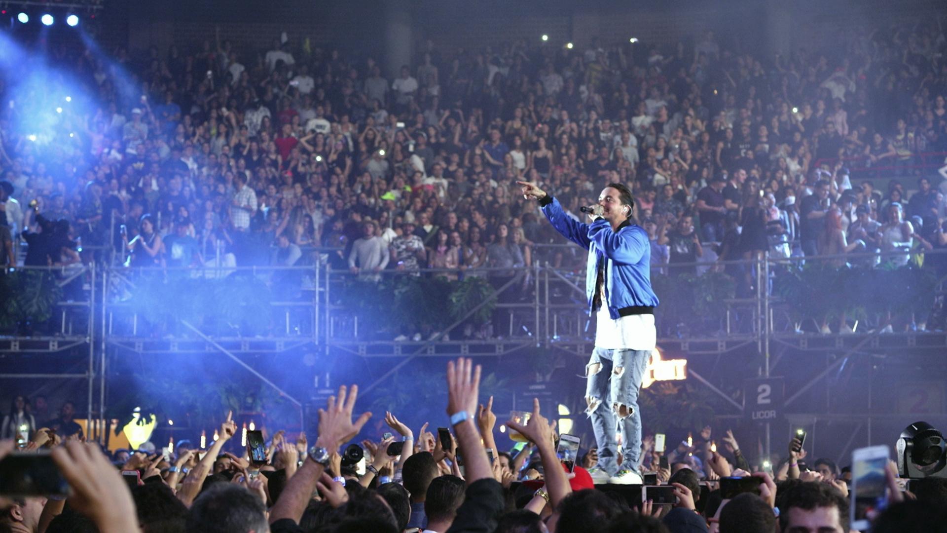 J Balvin in a blue jacket stands on a stage surrounded by a large, cheering crowd at a concert. Lights and smoke add to the vibrant atmosphere.