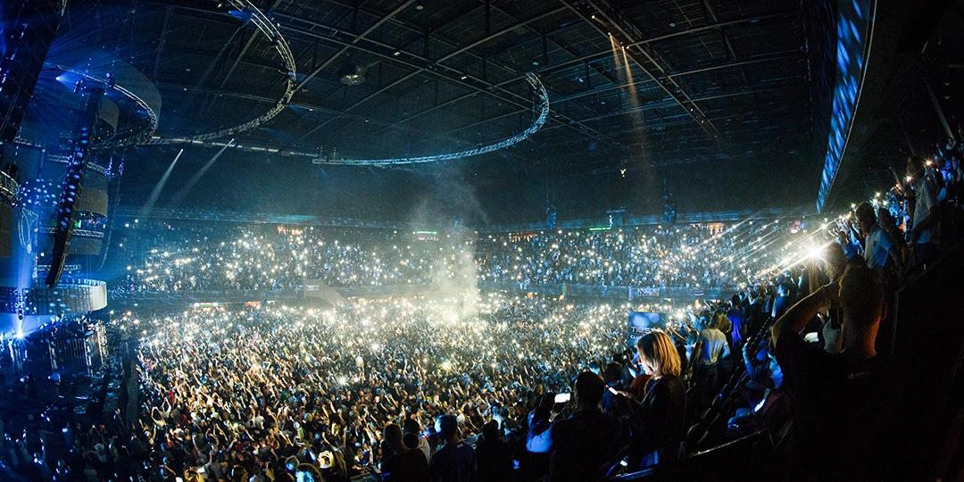 A large crowd at a concert venue, with many audience members using their phone flashlights, illuminating the dark arena. The stage and Tiësto are visible in the distance.