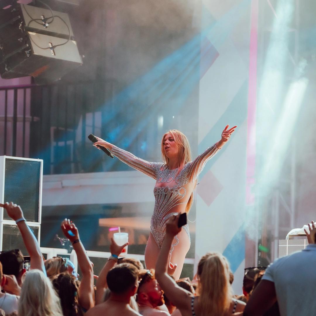 Becky Hill in a sparkling bodysuit performs energetically on stage in front of a lively crowd, holding a microphone and raising her arms. Multicolored lights and smoke add to the vibrant atmosphere.