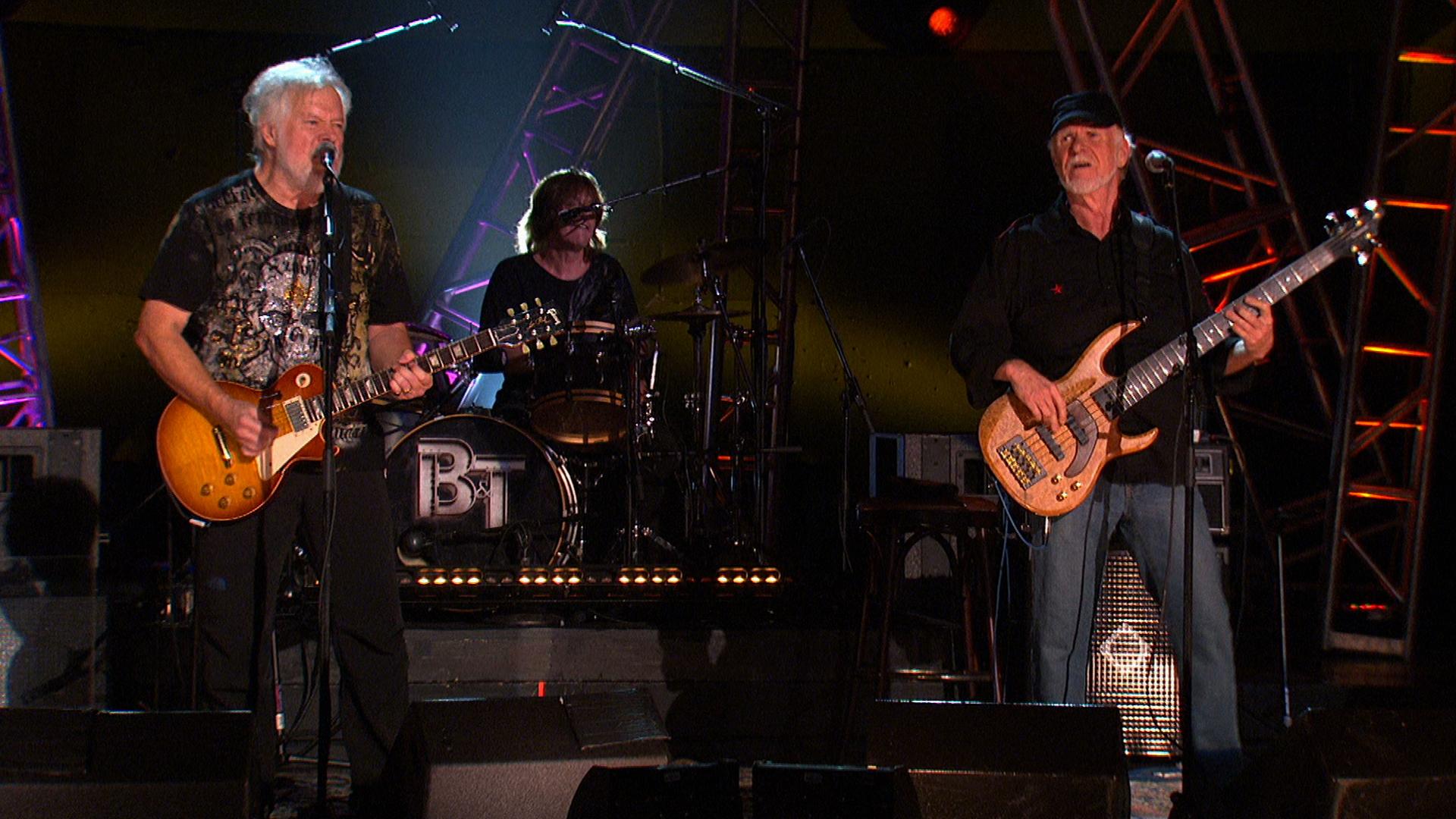 Bachman & Turner perform on a lit stage; one playing electric guitar, one playing drums, and one playing bass guitar. Stage structures and lights are visible in the background.