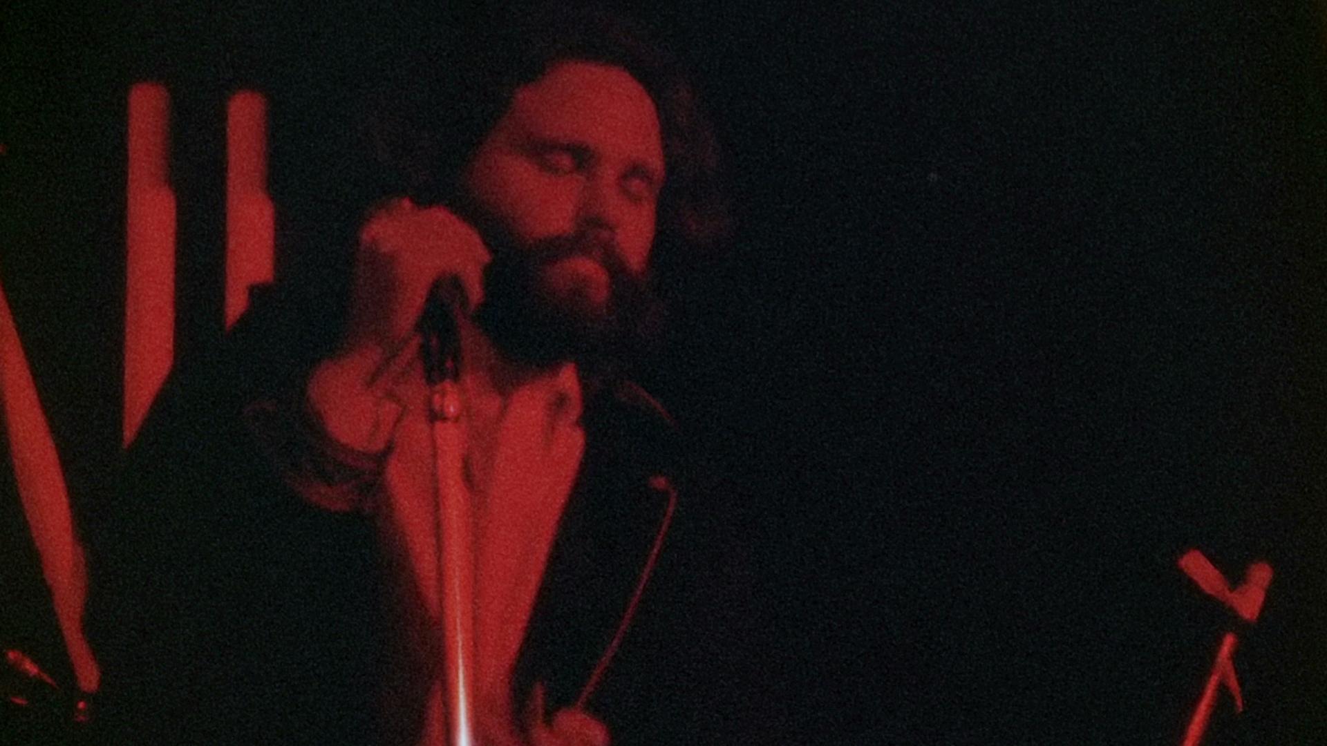 Jim Morrison stands on stage holding a microphone with eyes closed, illuminated by red lighting.