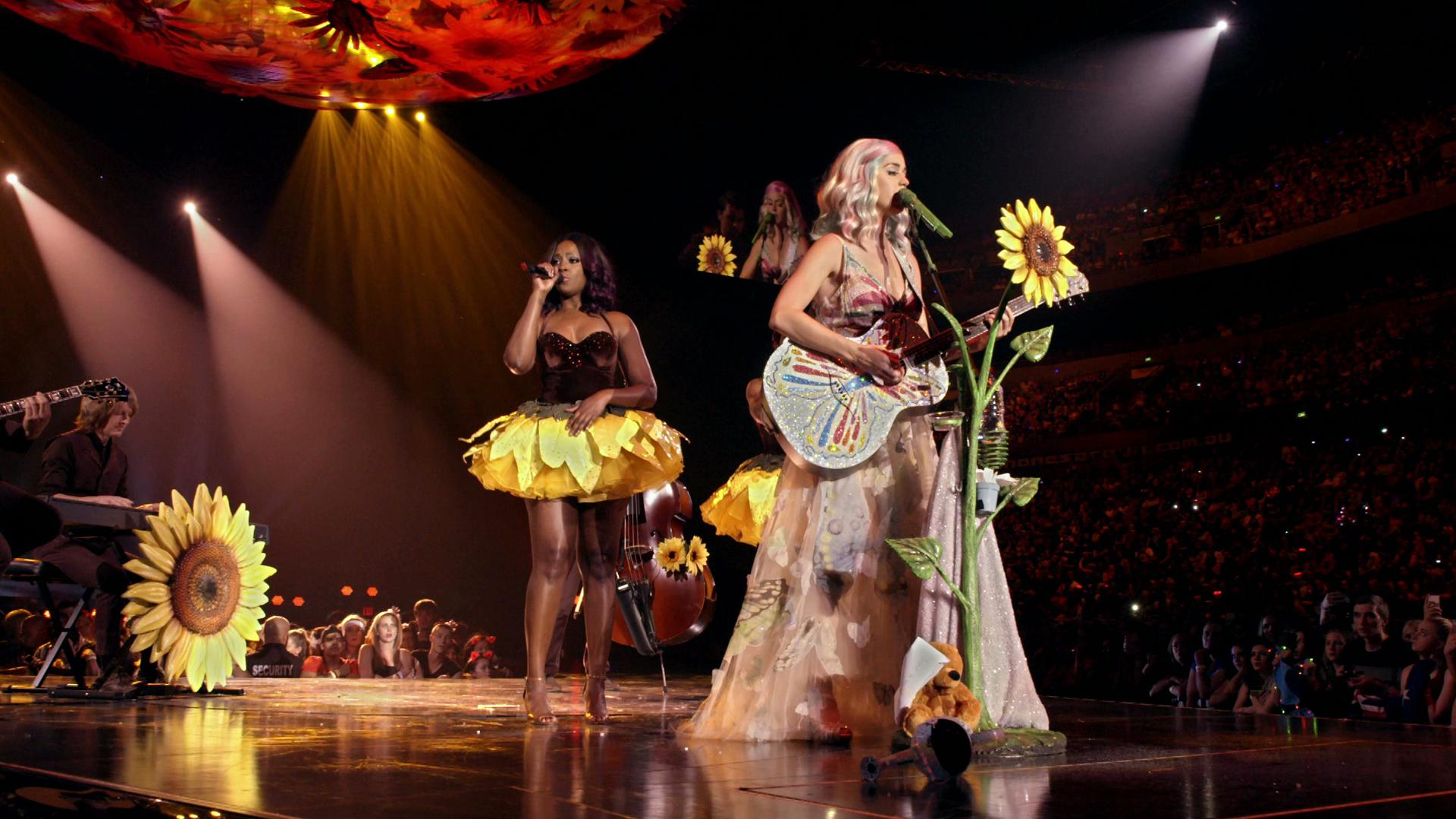 A performer in a flowing, floral-themed outfit plays a guitar adorned with sunflowers on a stage, accompanied by a singer in a sunflower skirt and cellist, under colorful overhead lighting.