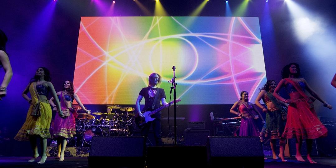 Steven Wilson plays a guitar on a stage with a colorful background, surrounded by dancers in colorful costumes. Stage lights shine in various hues.