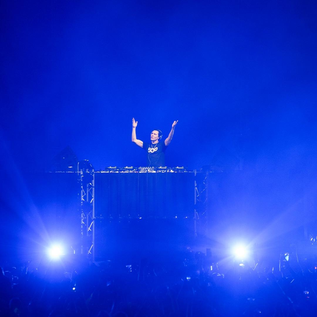 Tiësto stands behind a mixer on a stage with arms raised, illuminated by blue lights, in front of a cheering crowd.