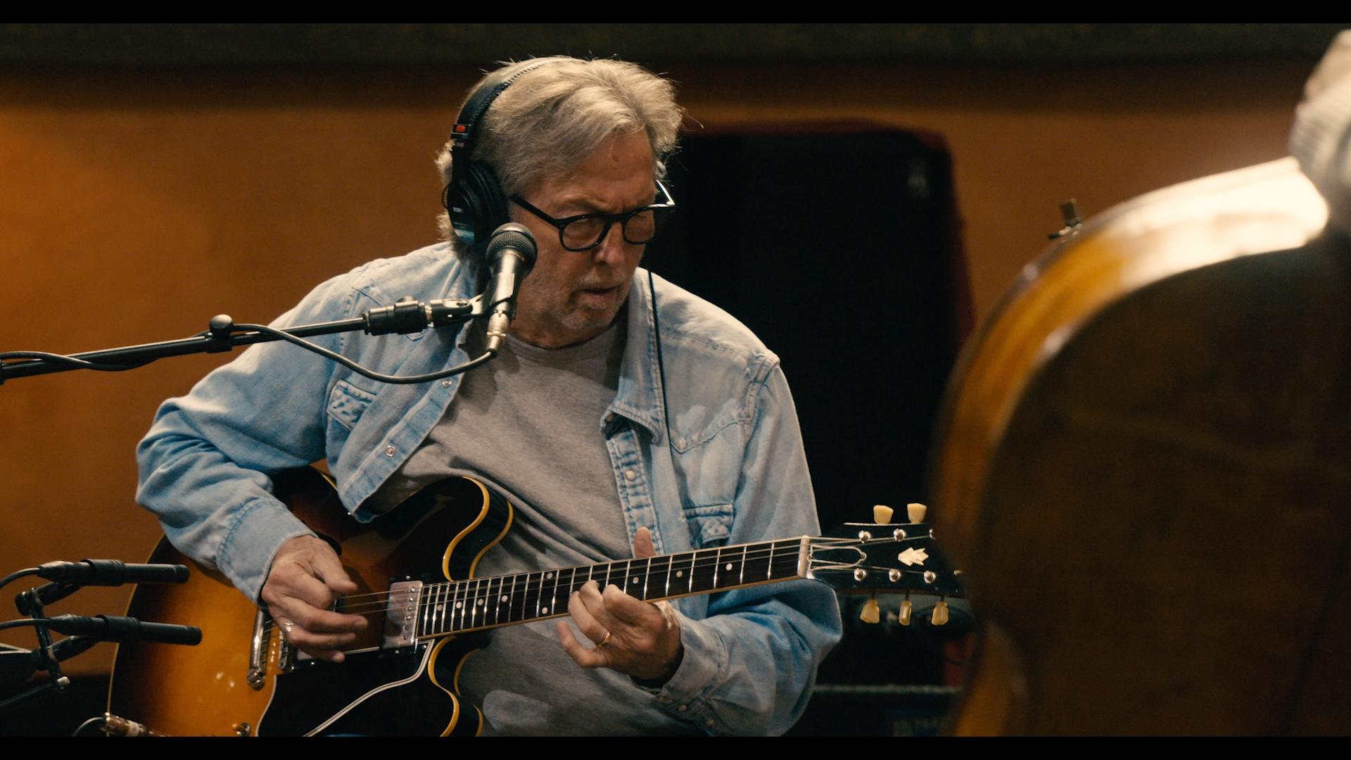 Eric Clapton playing an acoustic guitar and singing into a microphone while another person watches in a warmly lit room.