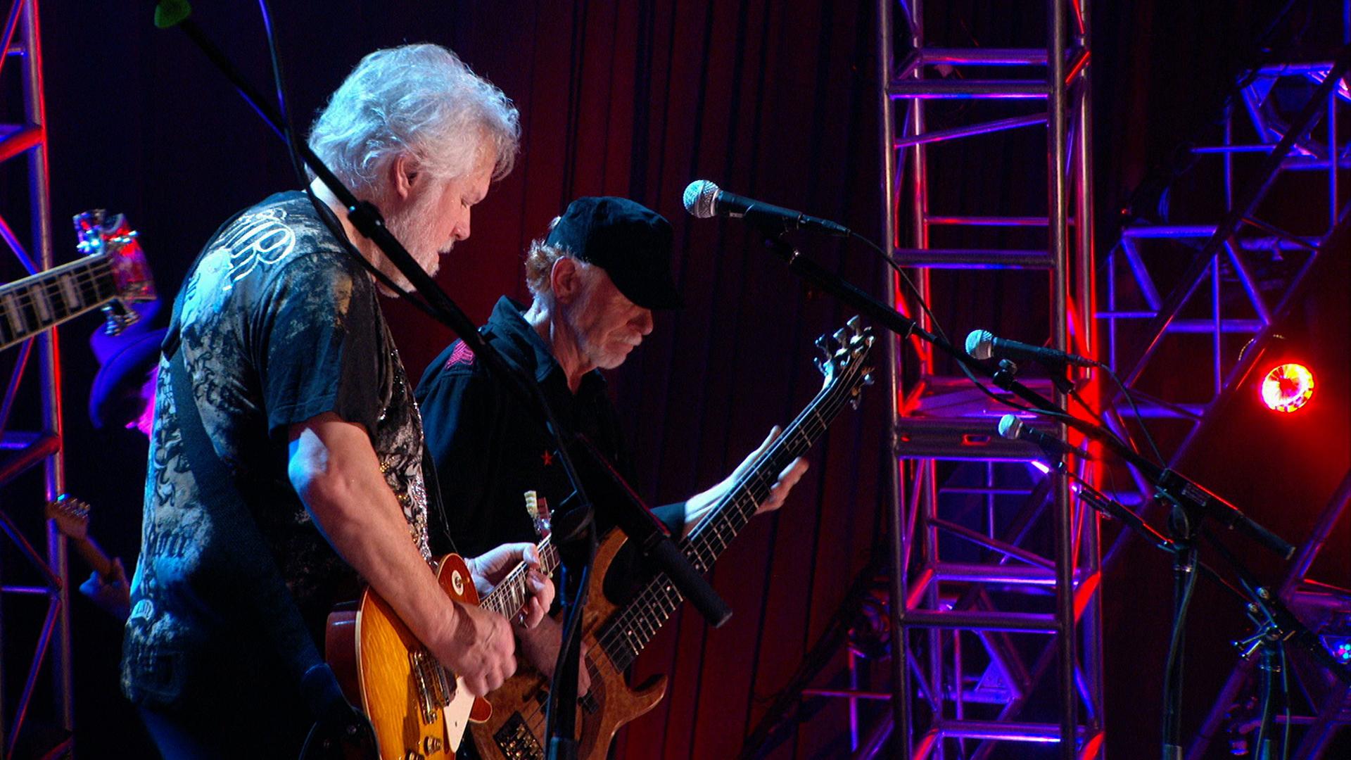 Bachman & Turner members playing guitars on a dimly lit stage with colorful lights and metal scaffolding in the background. The man on the left is playing an electric guitar, and the man on the right is playing a bass guitar.