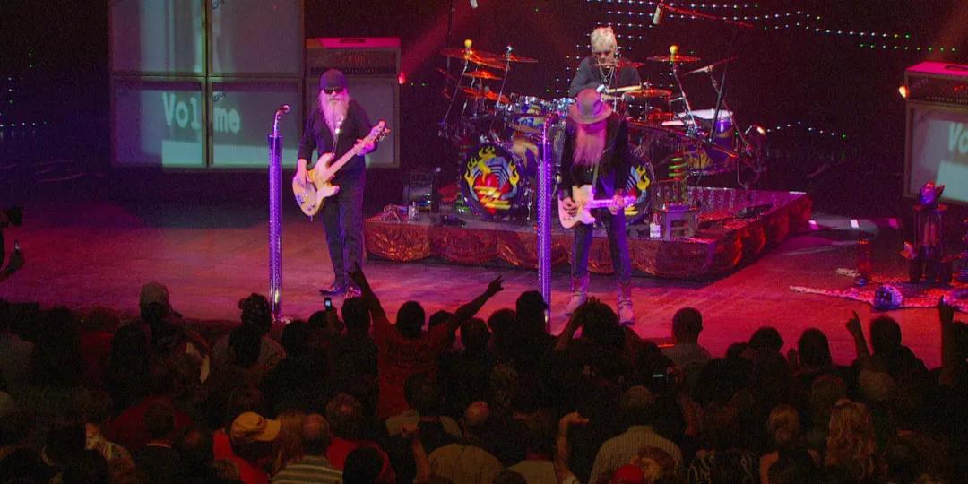 A three-member band performs on stage with guitars and drums, in front of a large audience. The stage is lit with colorful lights and decorated with amplifiers and a drum set.