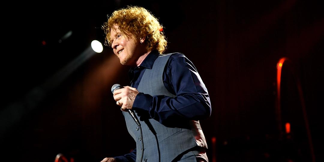 Simply Red with curly red hair sings into a microphone on stage, illuminated by focused lighting. They are wearing a blue shirt and gray vest.