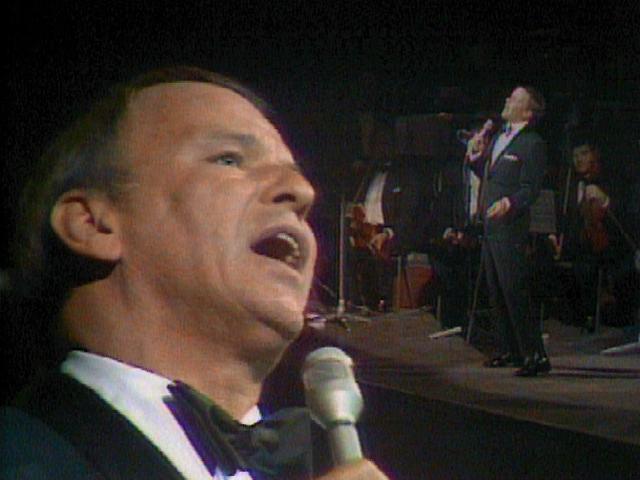 frank sinatra in a tuxedo sings into a microphone on stage with an orchestra in the background. A smaller image of him performing is superimposed over the larger image.