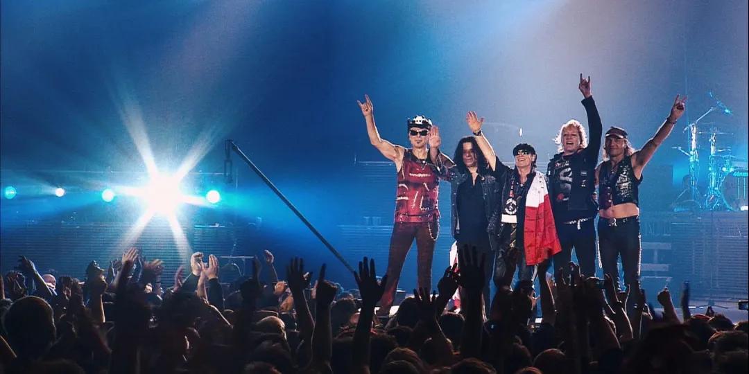 Scorpions stand on stage in front of a cheering crowd, raising their hands in a gesture as stage lights shine behind them.