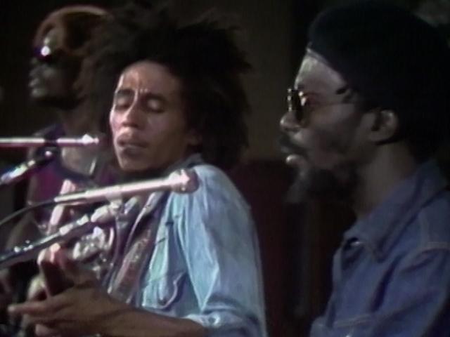 Bob Marley and two musicians perform on stage; one plays guitar and sings into a microphone. All are wearing sunglasses, and the central figure has a notable afro hairstyle.