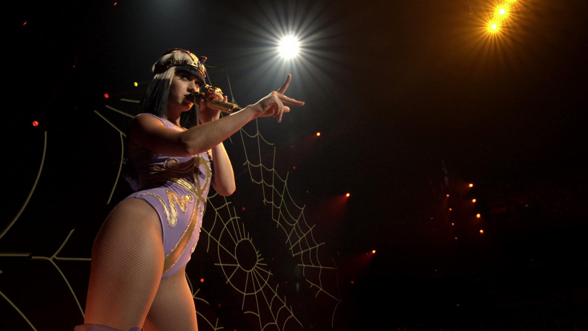 A performer is singing on stage under bright spotlights, wearing a detailed costume. A large spider web design is visible in the background.