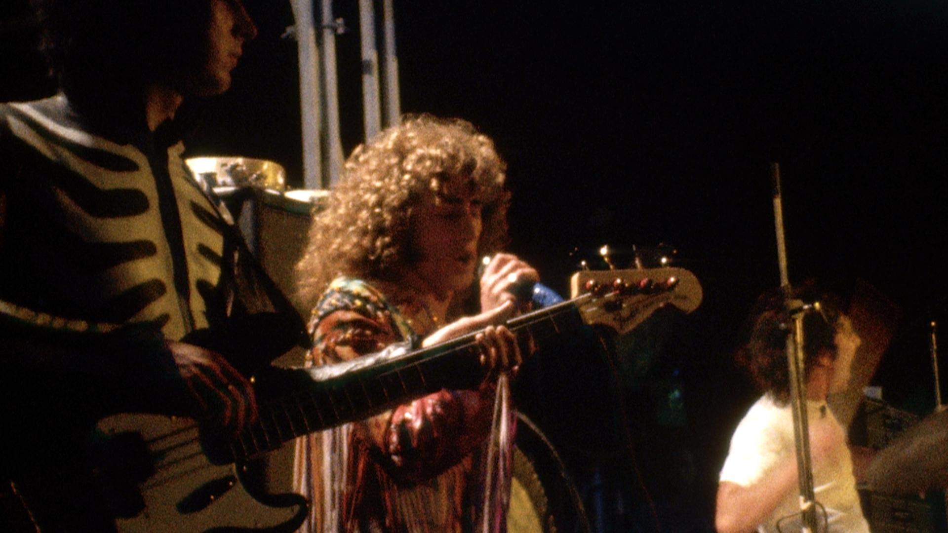  The Who playing an electric guitar on stage near a microphone stand, with amplifiers in the background.