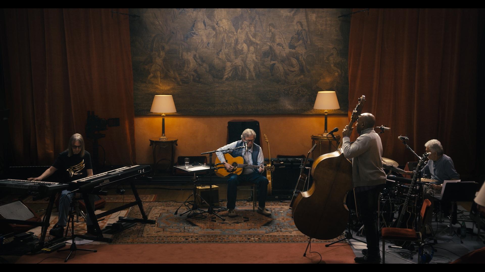 Eric Clapton playing in a dimly lit room. One plays keyboard, another acoustic guitar, another double bass, and the fourth one is on drums. The room has warm lighting, a tapestry, and various equipment.