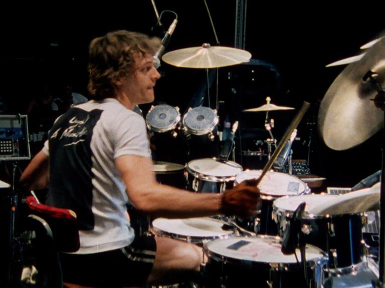 The Police drummer playing a drum set on stage, wearing a white and black shirt, holding drumsticks. Two microphones are positioned above the drum kit. The background includes various musical equipment.