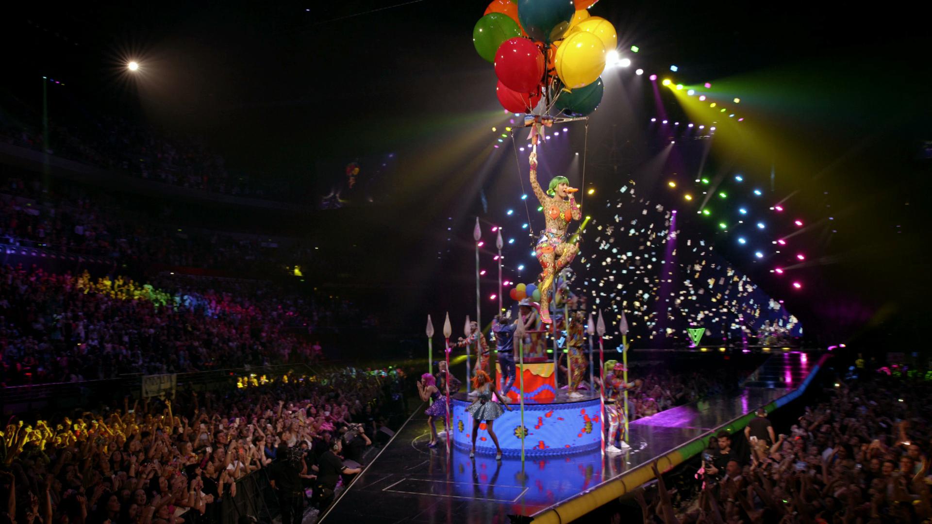 Katy Perry in a colorful costume is lifted by balloons above a stage during a lively concert with a cheering audience and vibrant lighting effects.