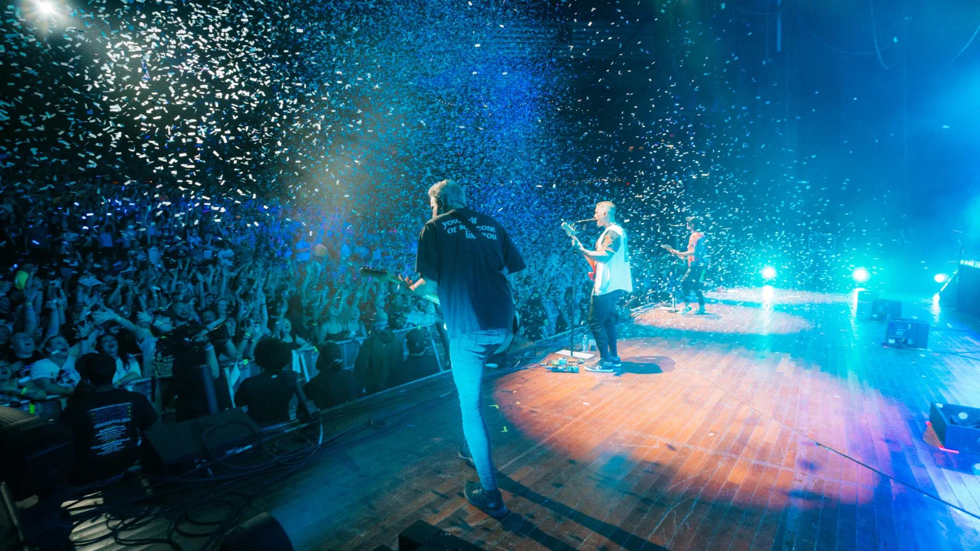 All Time Low performs on stage as confetti falls over a large, energetic audience in a concert hall with vivid blue and green lighting.