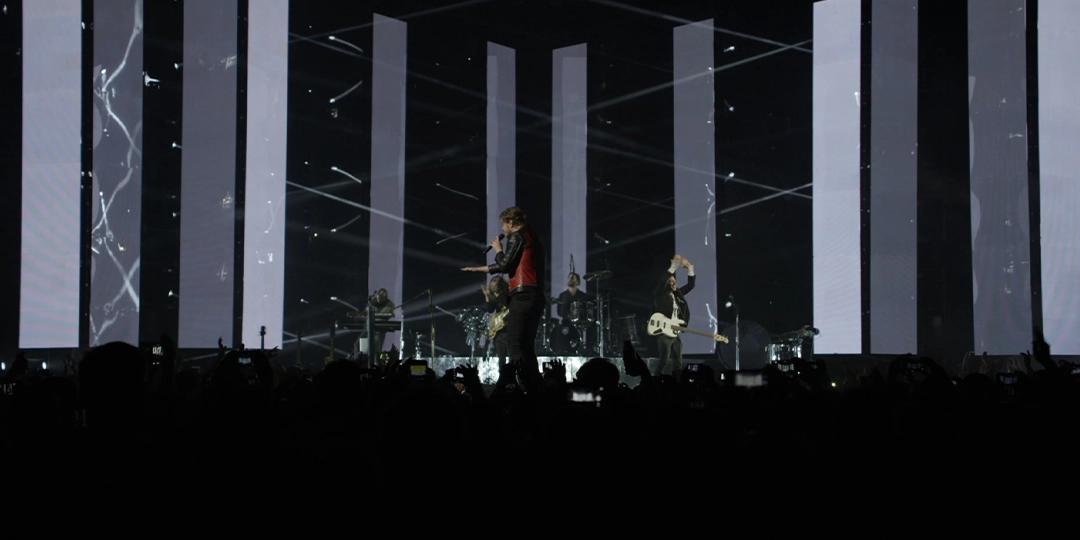 Imagine Dragons perform on stage with tall, vertical LED screens in the background displaying abstract patterns. The stage is lit with various spotlights and effects. The audience can be seen in the foreground.