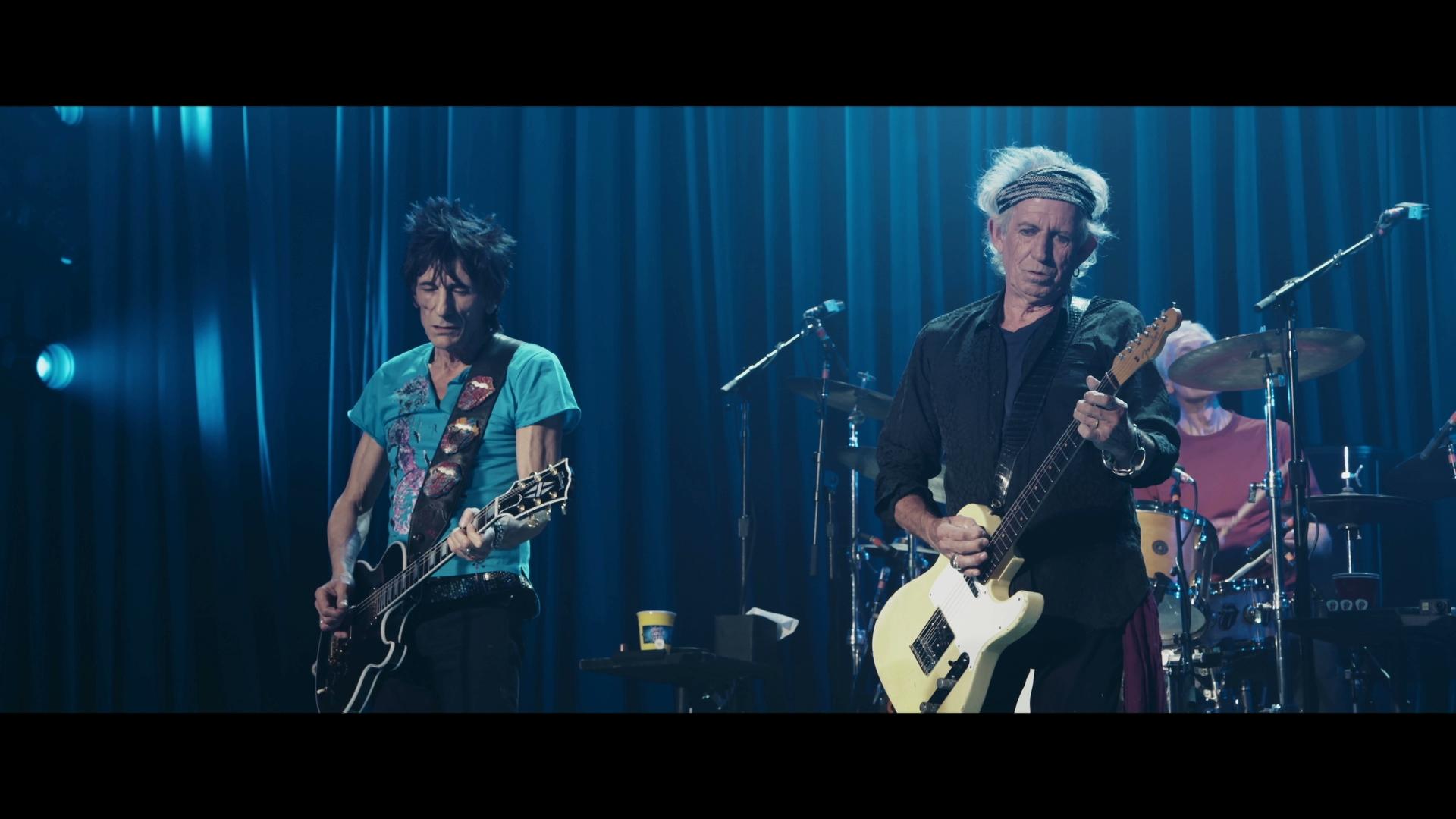 The Rolling Stones perform on a stage with blue lighting. The musician on the left plays a black electric guitar, wearing a blue shirt. The musician on the right plays a white electric guitar, wearing a black shirt.