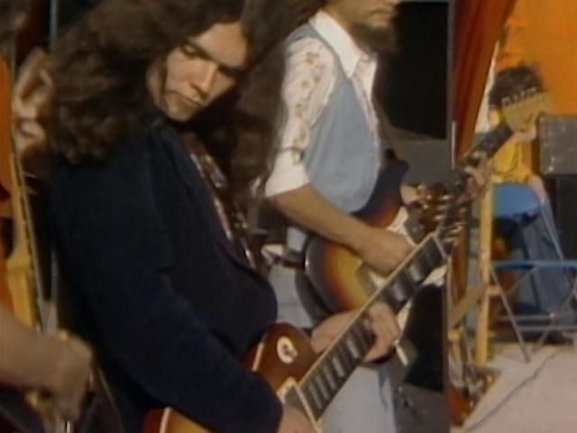 Lynyrd Skynyrd playing guitars, one in the foreground with long hair and a dark shirt, and the other in the background with a beard and wearing a blue vest over a patterned shirt.