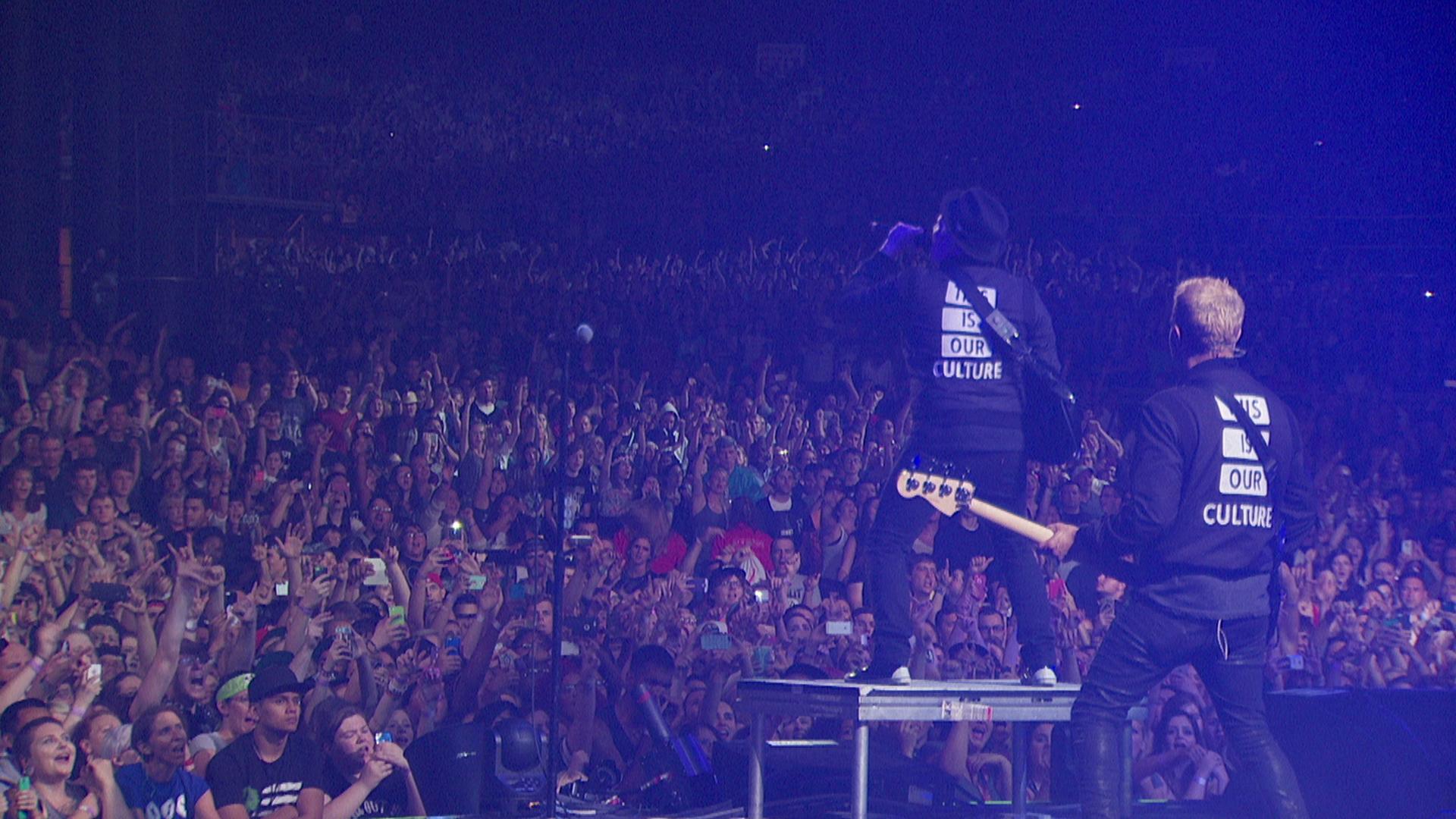 Fall Out Boy perform on stage in front of a large, cheering audience. One musician plays a bass guitar while the other stands on an elevated platform. Both wear black jackets with text on the back.