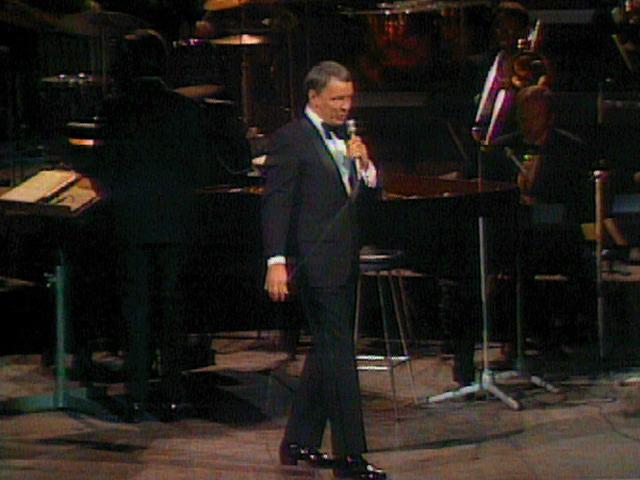 frank sinatra in a suit and bow tie performs on stage with a band in the background.