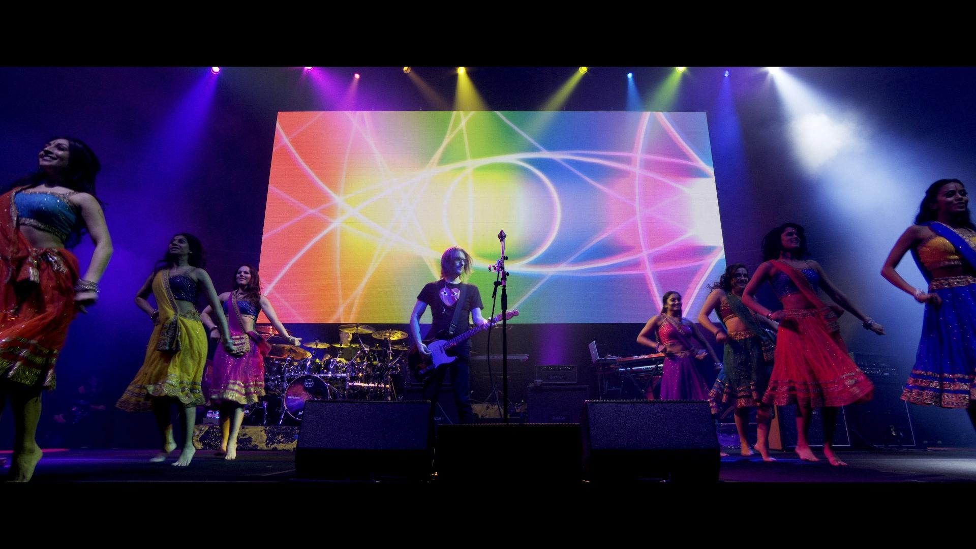 Steven Wilson plays guitar on stage surrounded by dancers in colorful costumes, with vibrant light patterns displayed on a large screen in the background.