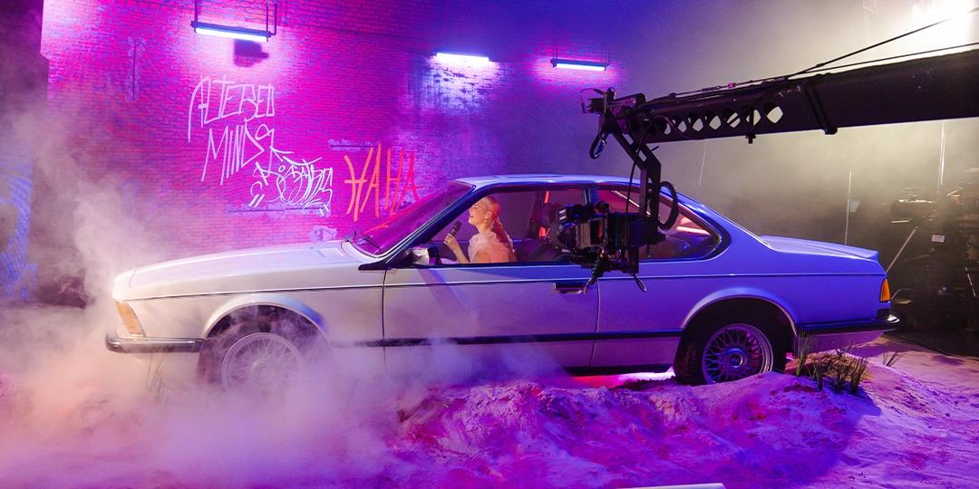 Anne-Marie is sitting in a white car on a set with purple lighting, a fog machine, and graffiti on the wall in the background.