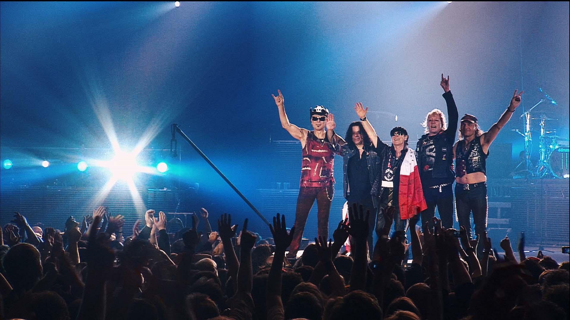 Scorpions wearing colorful outfits, stands on a stage waving to an audience while holding hands. A bright stage light shines in the background as the crowd raises their hands.