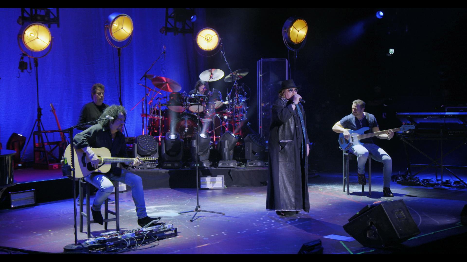 Toto performs on stage with blue lighting. The lead singer wears a black coat and hat, while other members play drums, guitar, and bass. Circular stage lights hang above them.