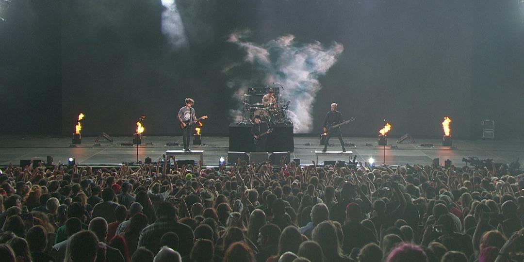 Fall out boy performs on stage with illuminated flames and a crowd in front of them. Smoke effects are visible in the background.