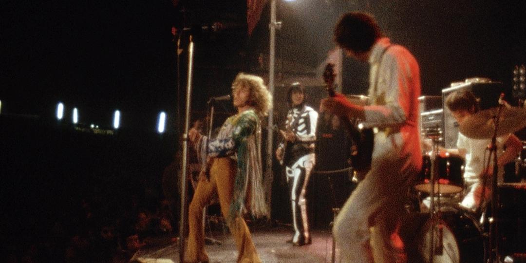 The Who performs energetically on stage under bright lights with one member dressed in a skeleton suit and others wearing colorful outfits.