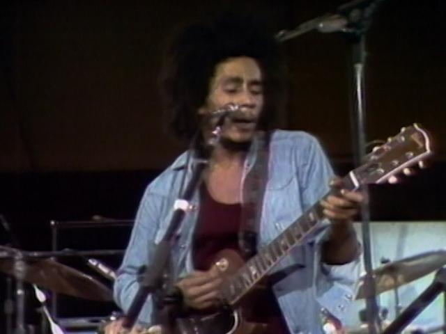 Bob Marley plays an electric guitar and sings into a microphone on stage, wearing a denim jacket over a red shirt.