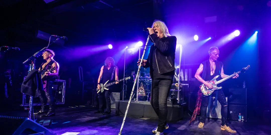 A rock band performs on stage with vibrant purple lighting. Four members play instruments, while one sings into a microphone.