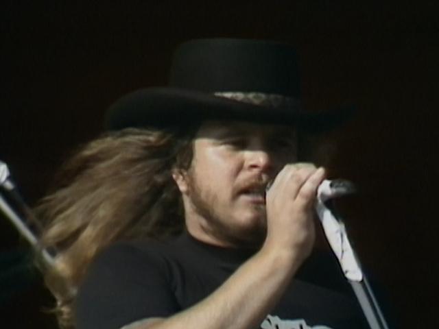 Lynyrd Skynyrd frontman with long hair and a beard wearing a black hat and shirt, holding a microphone close to their mouth, singing.