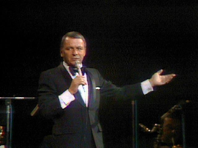 Frank Sinatra in a black suit is holding a microphone with his right hand and gesturing with his left hand while standing on stage.