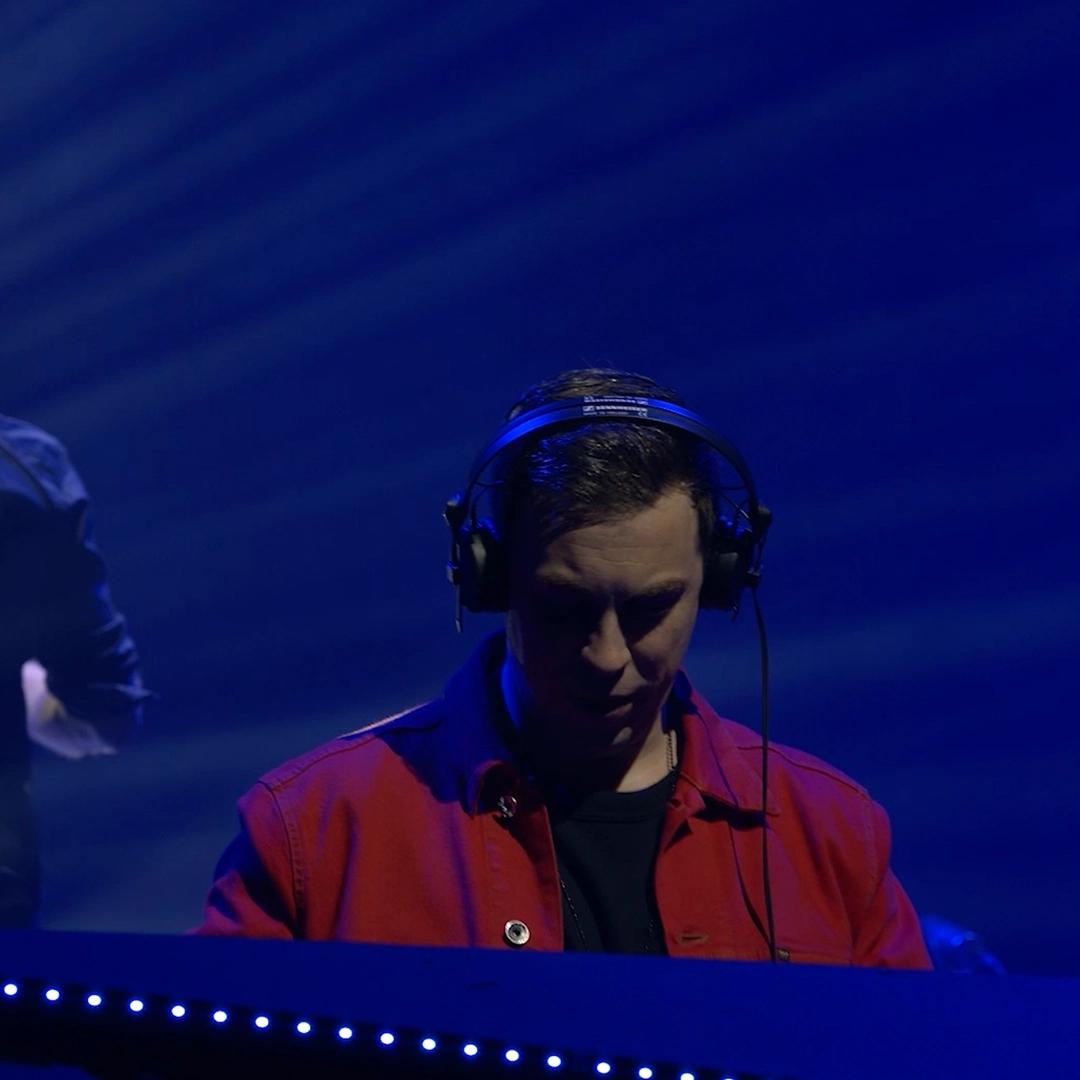 Hardwell wearing headphones and a red jacket performs on stage with blue lighting.