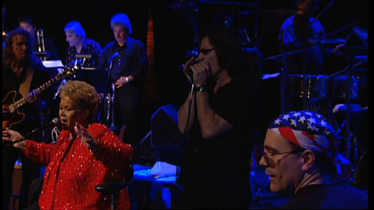 Etta James performs on stage, featuring a woman singing in a red outfit, a man playing harmonica, and musicians playing guitar and drums with a bandana-wearing man in the foreground.