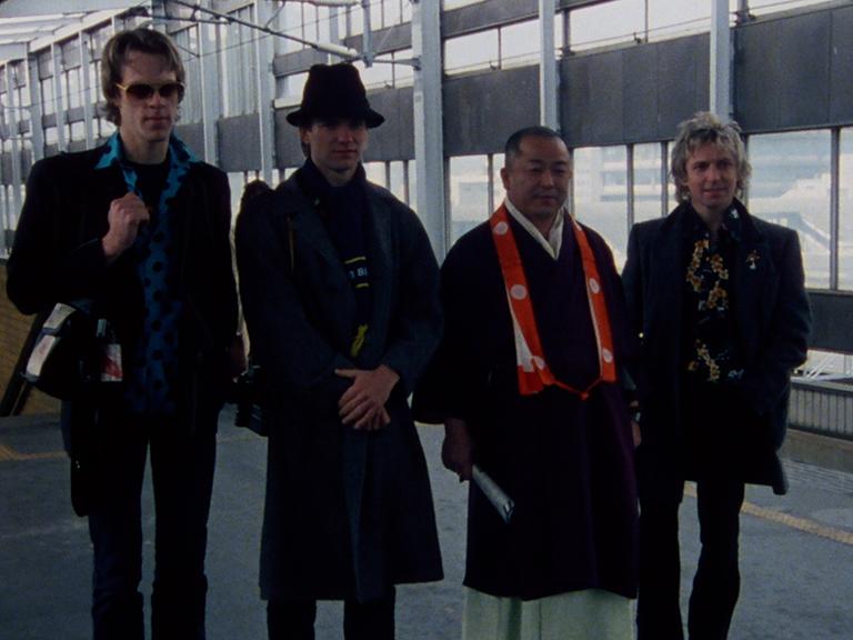 The Police stand together in a line, two casually dressed in sunglasses, one in a robe, and one in a dark coat and hat, in an indoor setting with large windows.