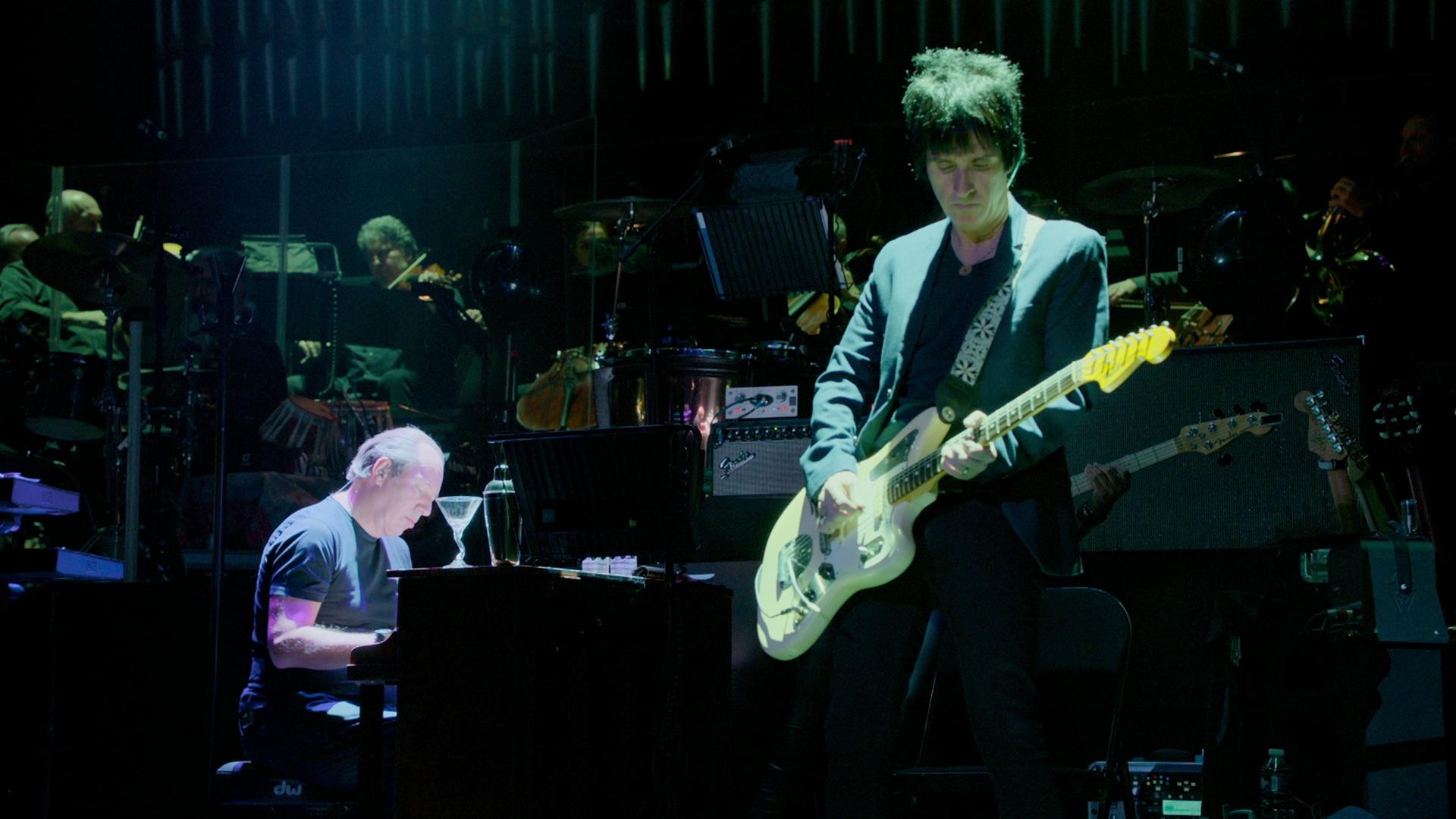 Hans Zimmer and Johnny Marr performing on stage, one playing the keyboard and the other playing an electric guitar. In the background, other musicians can be seen playing various instruments.