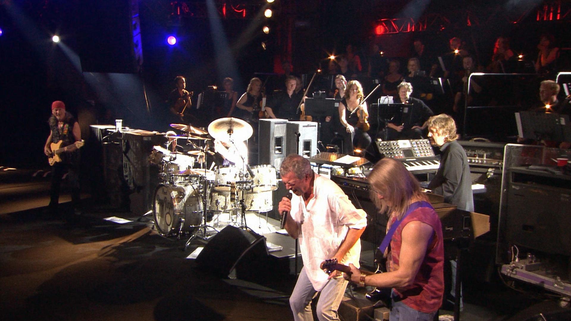 Deep Purple performs on stage with various instruments; a keyboardist, guitarist, bassist, drummer, and vocalist. An orchestra is seated in the background, with musicians playing string instruments.