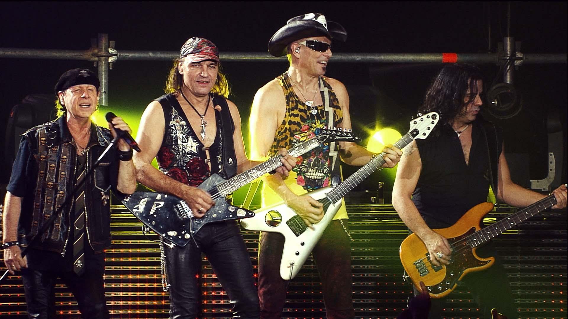 Scorpions perform energetically on stage, each playing instruments: two electric guitars, a bass guitar, and one singing with a microphone, with bright stage lights illuminating the background.